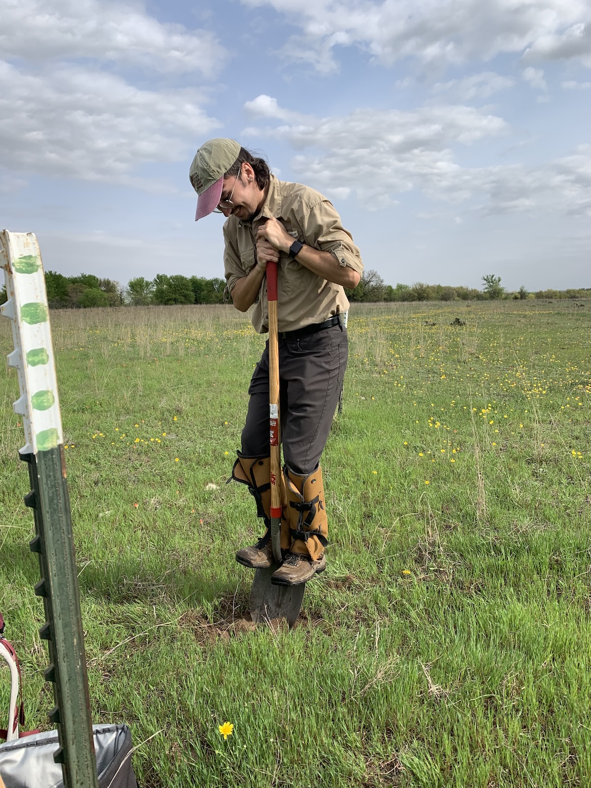 Young man standing on a shovel that is partly into the ground, in the middle of a field