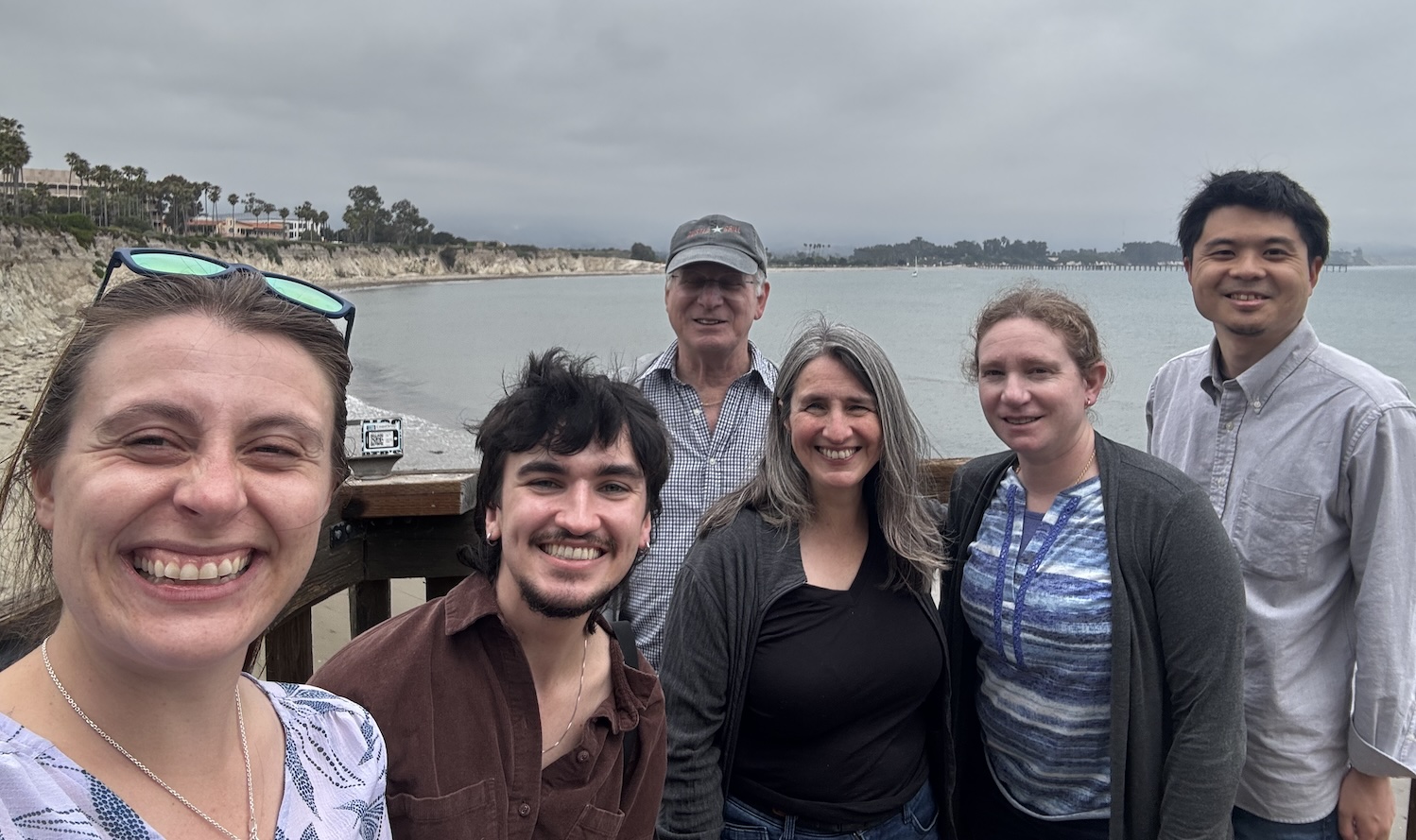 Six people are gathered in front of the camera, with the beach and a cloudy sky in the background