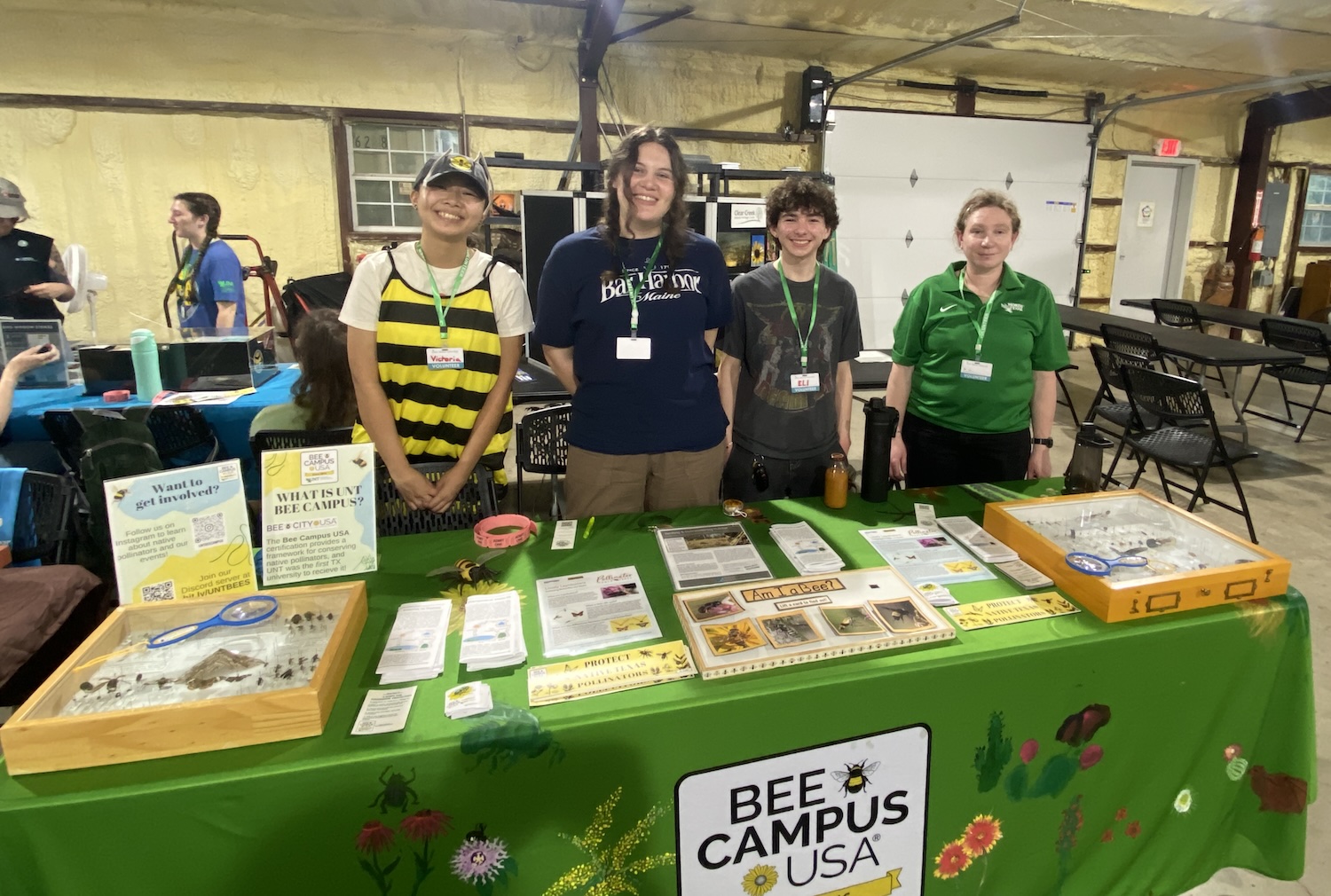 Four people, one wearing a bee costume, standing behind a table that contains displays related to pollinators